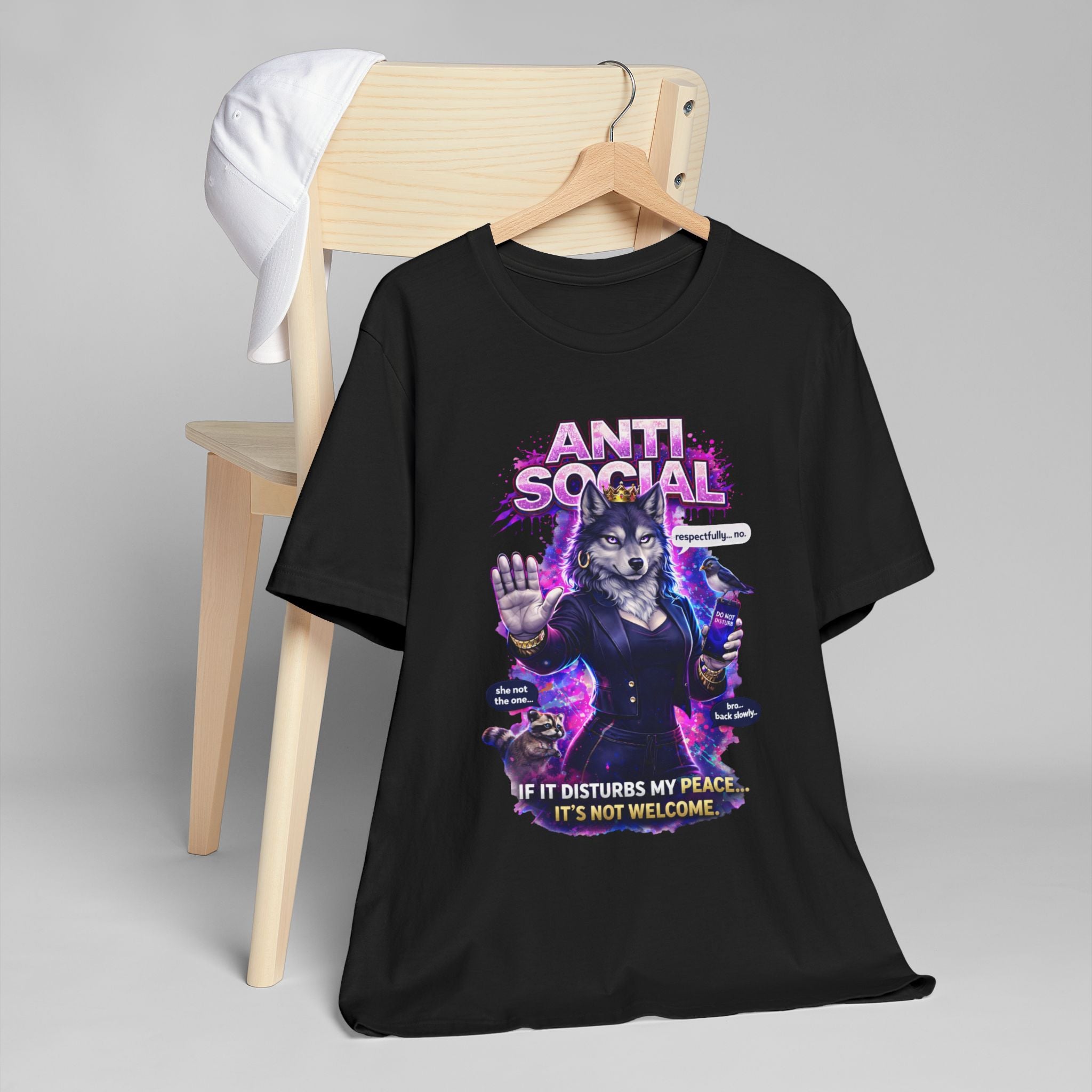 The Anti Social She-Wolf T-Shirt — Funny Introvert Graphic Tee features a black design with a wolf in a suit and the caption "If it disturbs my peace... it's not welcome," shown draped over a wooden chair with a white cap—perfect for bold personalities.