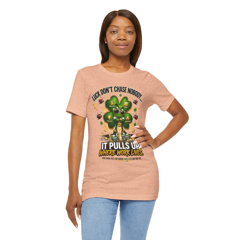 A woman poses with one hand on her hip, wearing the TimeWarp Drip Lucky Clover "Pulls Up Lives" Graphic T-Shirt in peach (sizes S-3XL), featuring a cartoon four-leaf clover, bees, and the phrase: Luck don’t chase nobody. It pulls up where work lives.