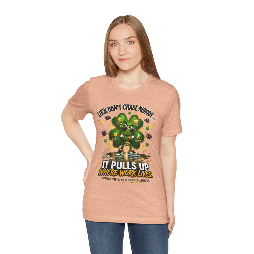 A woman with straight light brown hair wears the TimeWarp Drip Lucky Clover "Pulls Up Lives" Graphic T-Shirt in peach (sizes S–3XL) featuring a four-leaf clover, dollar signs, ladybugs, and the phrase: Luck don’t chase nobody—it pulls up where work lives.