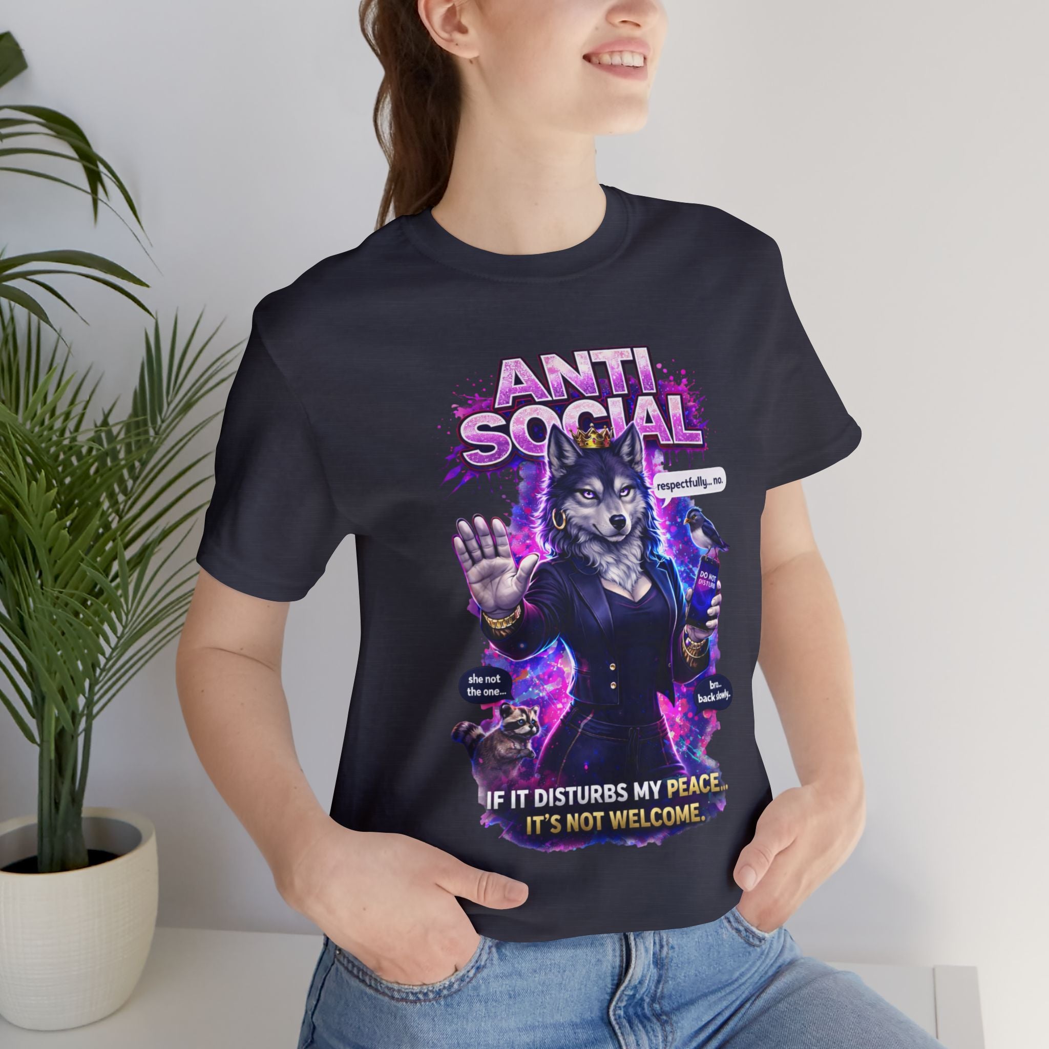 A smiling woman wears the Anti Social She-Wolf T-Shirt—Funny Introvert Graphic Tee, showing an anthropomorphic wolf in a suit with "ANTI-SOCIAL. If it disturbs my peace, it's not welcome." Green plants can be seen in the background.