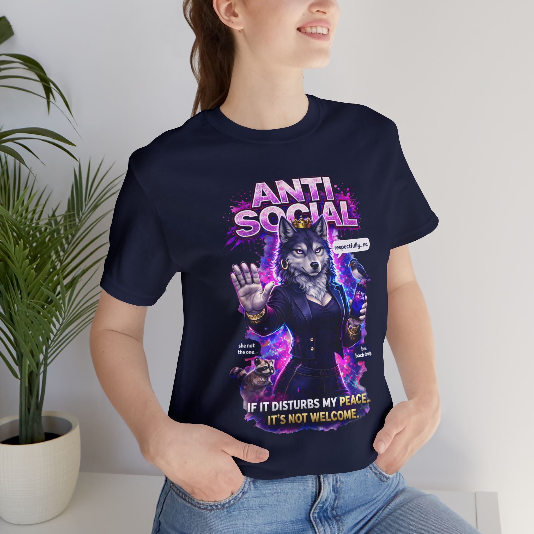 A smiling person wears the Anti Social She-Wolf T-Shirt — Funny Introvert Graphic Tee in navy blue, showing a wolf in a suit with raised hand, bright purple accents, and the text: If it disturbs my peace, it's not welcome. A potted plant sits behind them.