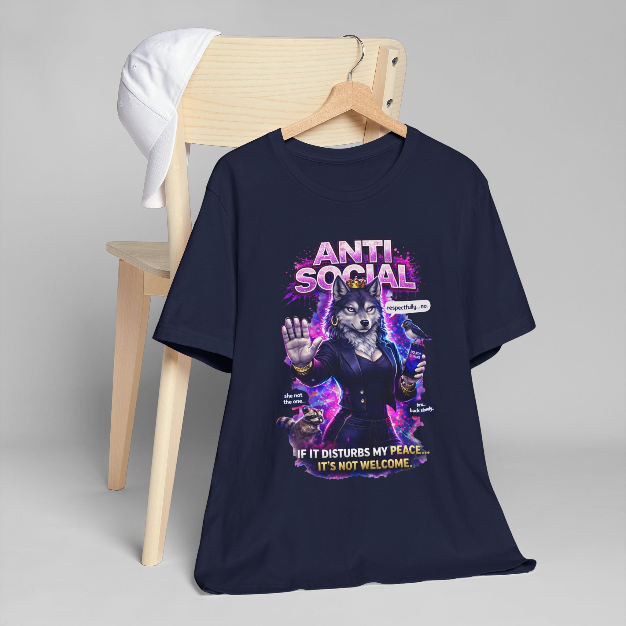 A navy blue Anti Social She-Wolf T-Shirt—Funny Introvert Graphic Tee—draped over a wooden chair with a white cap. Features a grumpy, suited she-wolf raising her hand and text: “If it disturbs my peace… it’s not welcome.”.