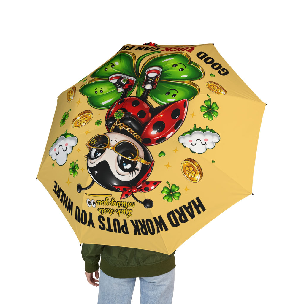 A person holds the Cute Lucky Ladybug Foldable (Yellow) Umbrella, featuring a cartoon ladybug in sunglasses, four-leaf clover, smiling clouds, gold coins, and the canopy text: HARD WORK PUTS YOU WHERE GOOD LUCK CAN FIND YOU.