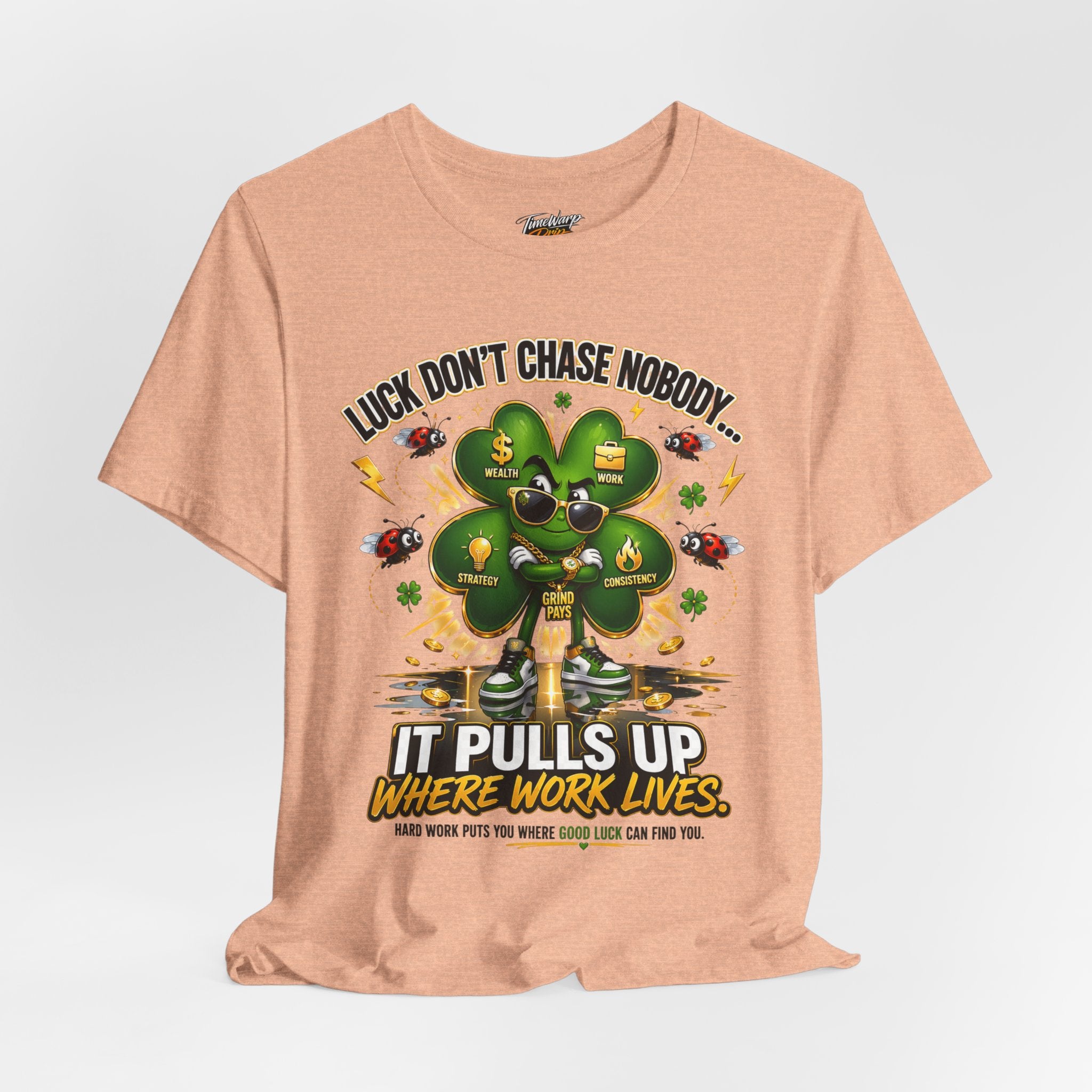 The TimeWarp Drip Lucky Clover "Pulls Up Lives" Graphic T-shirt, in peach (S-3XL), features a clover with sunglasses and gold chains, ladybugs, dollar signs, confetti, and the text: “Luck Don’t Chase Nobody… It Pulls Up Where Work Lives.”.