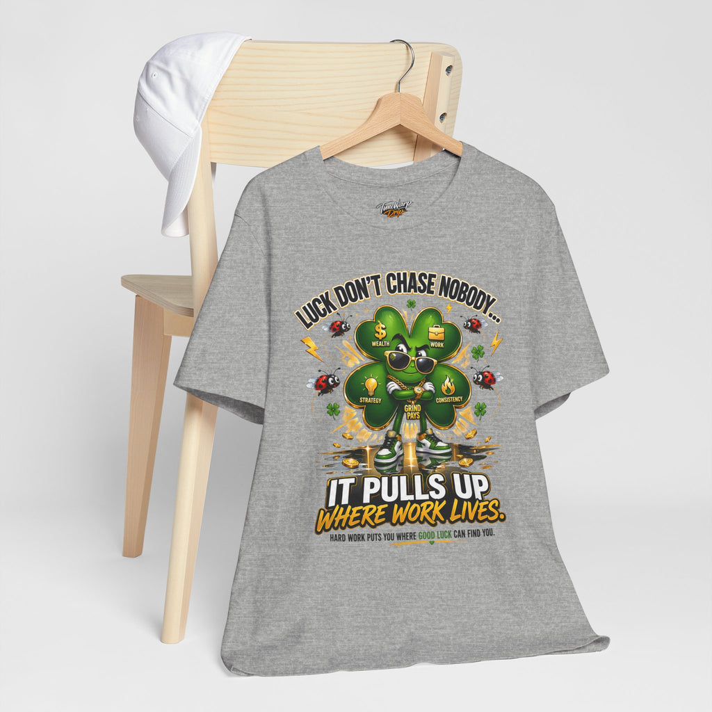 The TimeWarp Drip Lucky Clover "Pulls Up Lives" Graphic T-Shirt, available in gray (sizes S–3XL) and featuring a four-leaf clover with motivational text, hangs on a wooden chair with a white cap resting on the back.