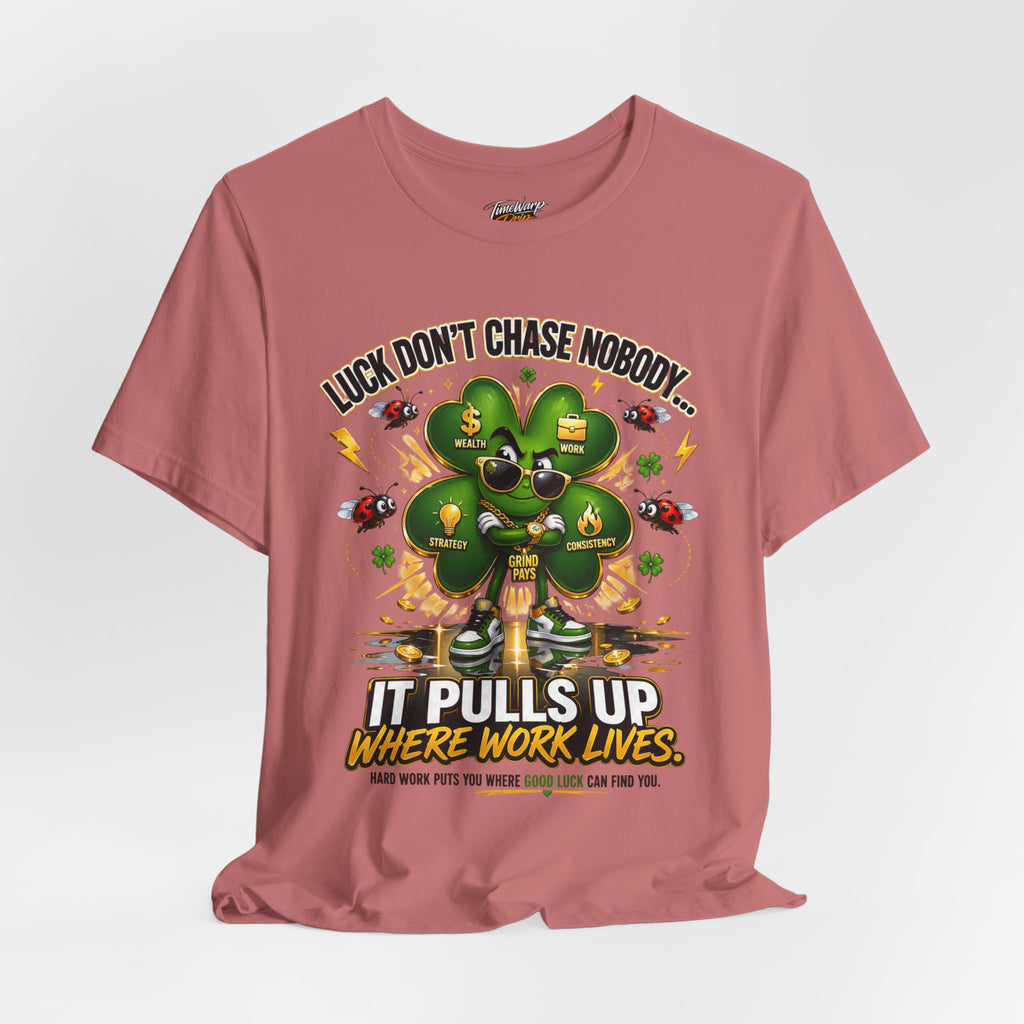 The Lucky Clover "Pulls Up Lives" Graphic T-Shirt in pink (sizes S–3XL) showcases a clover with sunglasses and gold chains, surrounded by wealth icons, plus the text: Luck don’t chase nobody… it pulls up where work lives.