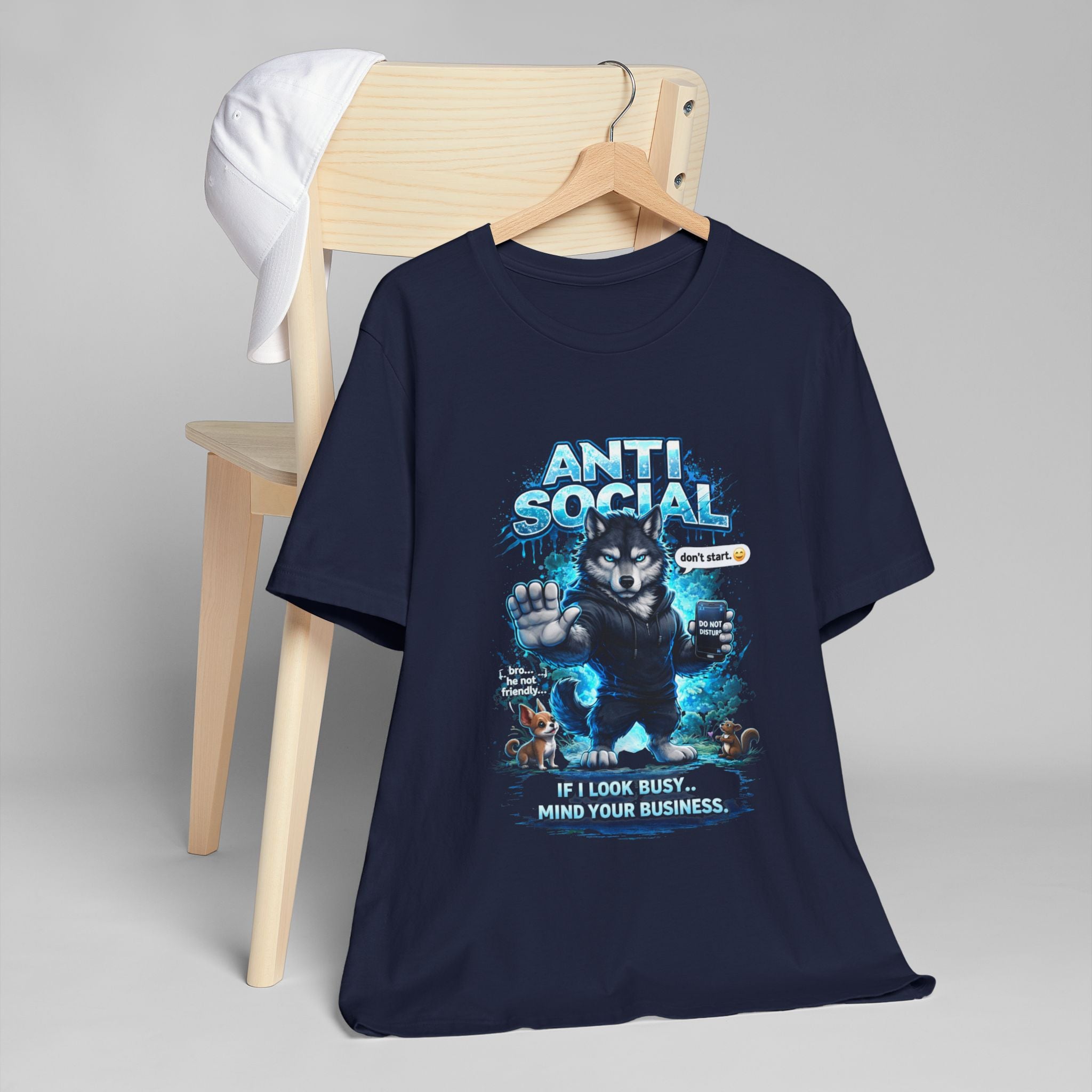 The Anti Social Wolf T-Shirt — Funny Introvert Graphic Tee by TimeWarp Drip features a grumpy wolf with smartphone and "ANTI SOCIAL" text, making a bold streetwear statement. Displayed draped on a wooden chair with a white cap on top.