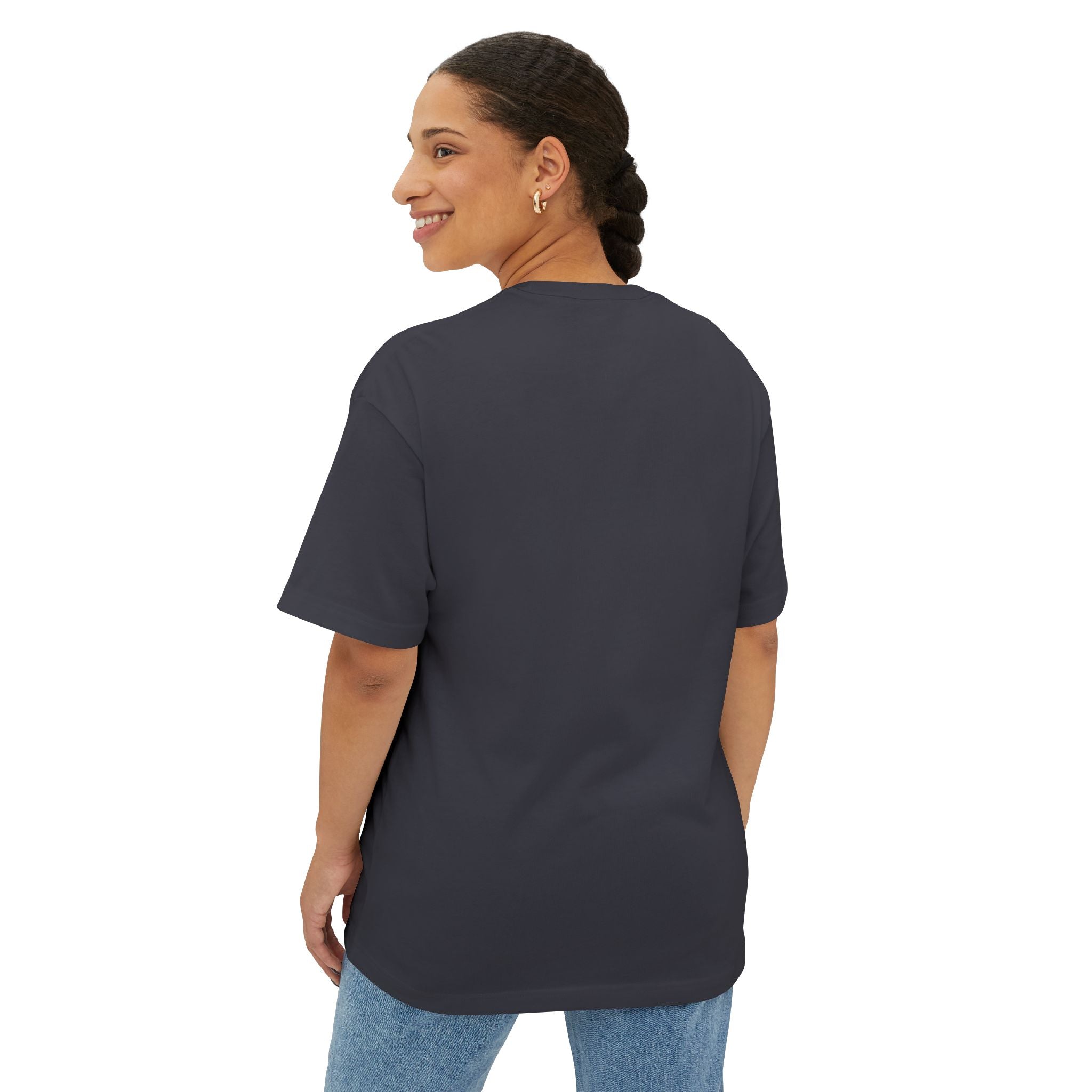 A woman with braided hair, wearing the Crowned In Every Era Unisex Oversized Boxy Tee in plain dark gray and light blue jeans, stands facing away while smiling and looking over her shoulder.