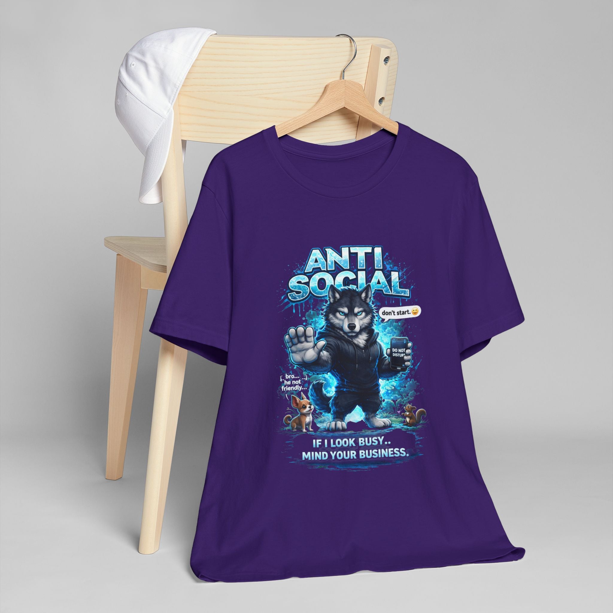 The Timewarp Drip Anti Social Wolf T-Shirt — Funny Introvert Graphic Tee in purple, with a grumpy wolf holding a phone and "ANTI SOCIAL. If I look busy… mind your business." text, is draped over a wooden chair topped with a white cap.
