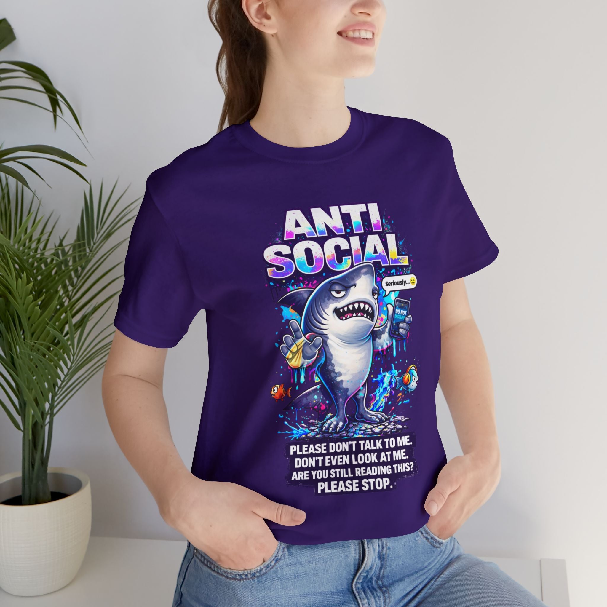 A smiling person wears the Anti Social Shark T-Shirt — Funny Introvert Graphic Tee, a unisex cotton shirt featuring a cartoon shark with snacks and drinks. The shirt reads "ANTI SOCIAL" alongside playful warnings not to talk or look.
