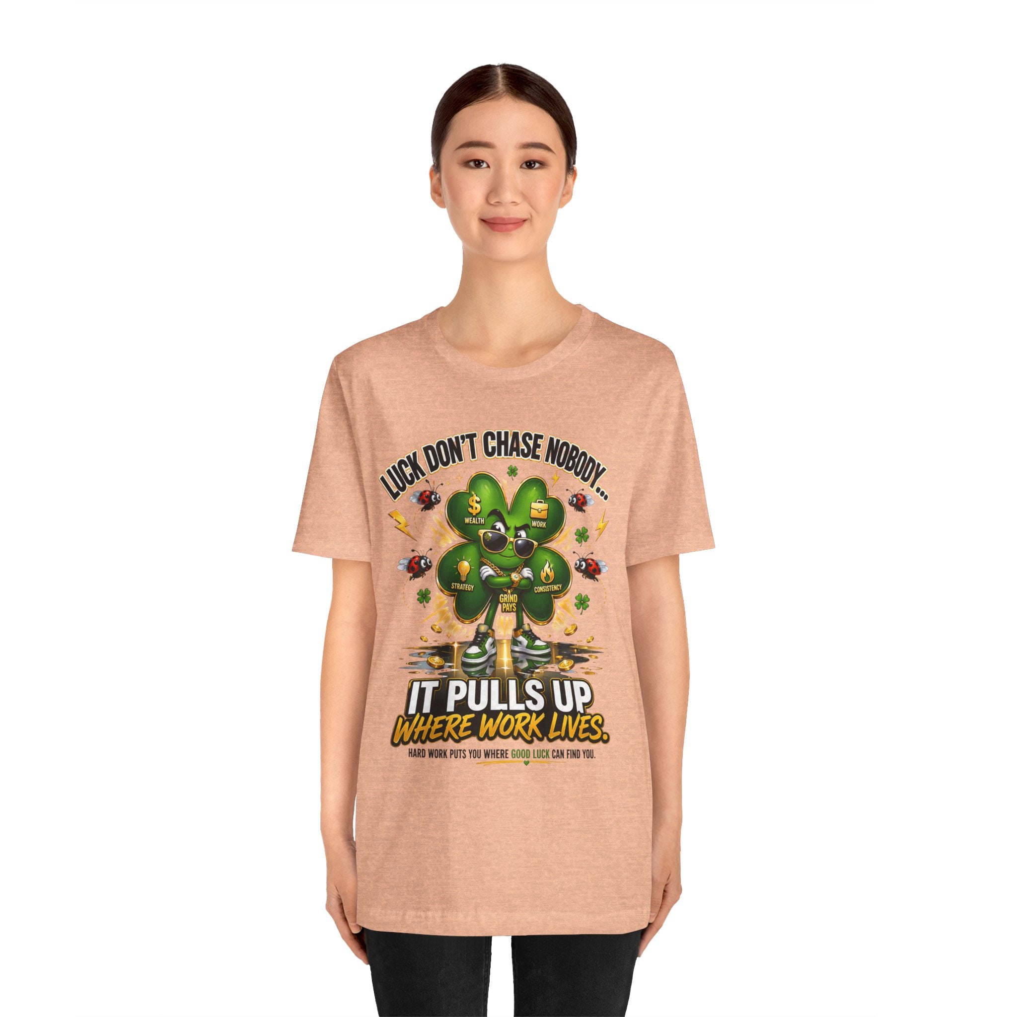 A young woman smiles at the camera wearing TimeWarp Drip's Lucky Clover "Pulls Up Lives" Graphic T-Shirt, available in sizes S-3XL. The light brown tee features a cartoon four-leaf clover and the text: Luck doesn’t chase nobody. It pulls up where work lives.