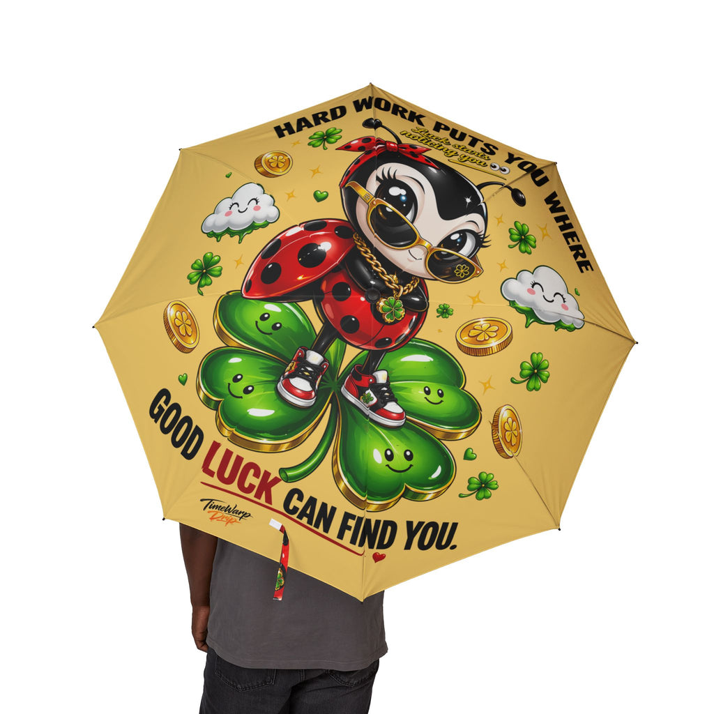 A person holds the Cute Lucky Ladybug Foldable (Yellow) Umbrella, featuring a cartoon ladybug in sneakers and sunglasses on a four-leaf clover with gold coins, clouds, and the text "Hard work puts you where good luck can find you.