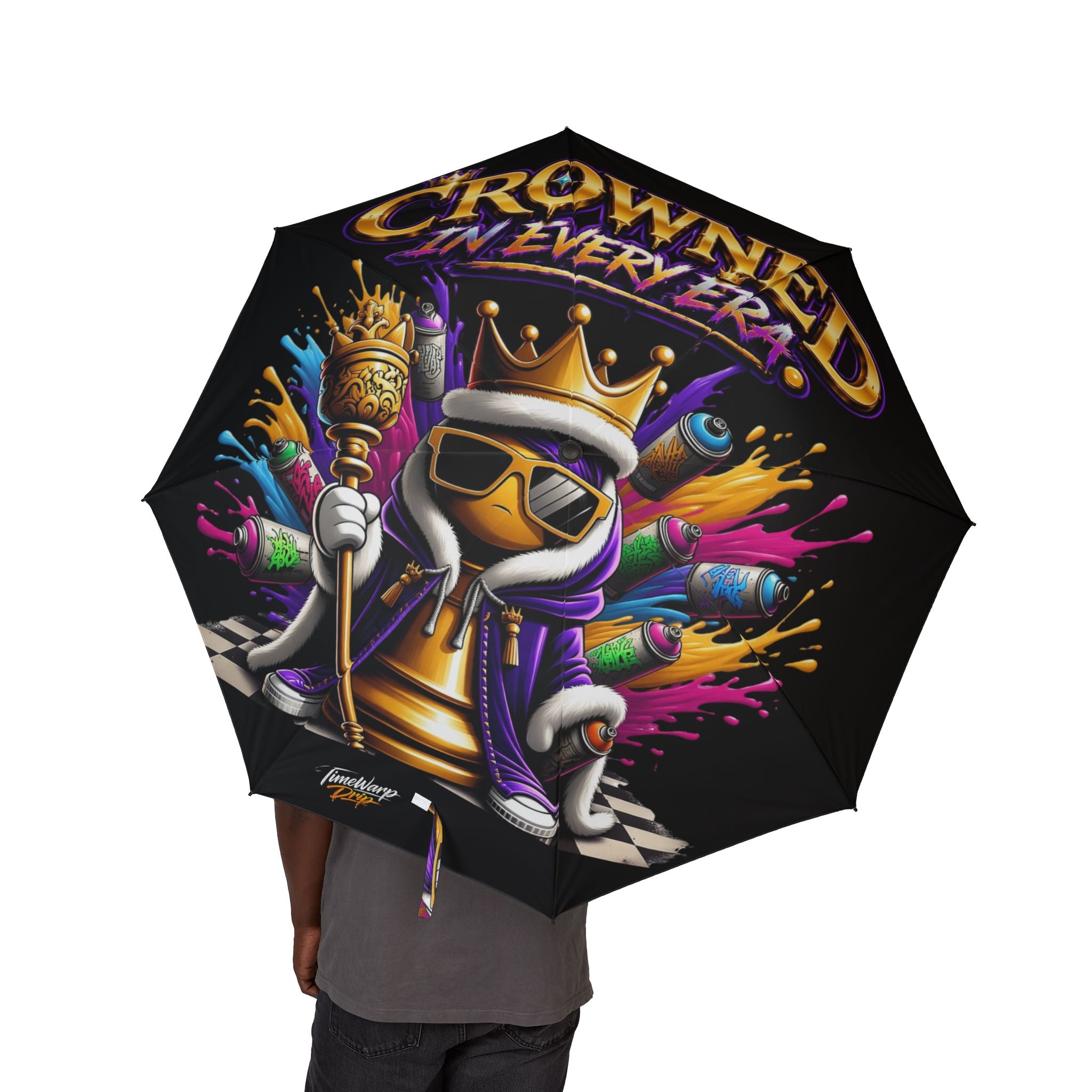 A person holds the Bold "Crowned In Every Era" Design — (Black) Umbrella (Semi-Automatic), featuring a cartoon chess king in a hoodie, crown, and sunglasses, with spray paint cans and the phrase "Crowned in Every Era.