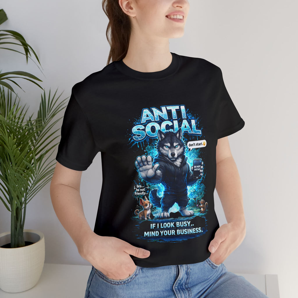 A woman stands indoors by a green potted plant, smiling with one hand in her jeans pocket, wearing Timewarp Drip’s Anti Social Wolf T-Shirt—Funny Introvert Graphic Tee featuring a grumpy wolf and the text “If I look busy, mind your business.”.