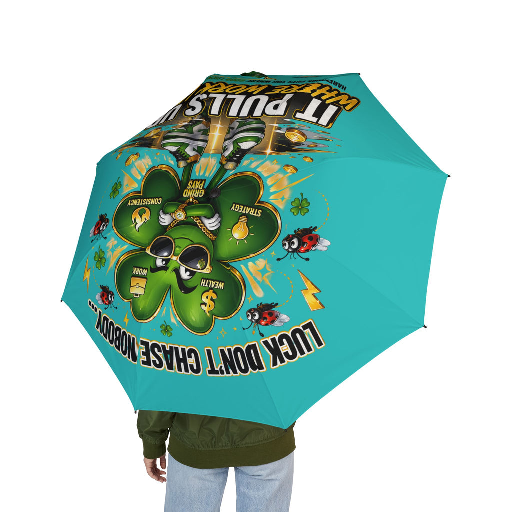 A person in a green jacket holds the Lucky Clover "Pulls Up Lives" teal foldable umbrella, featuring clovers, cartoon bees, dollar signs, and the phrases IT PULLS IN WHAT’S YOURS and LUCK DON’T CHASE NOBODY.